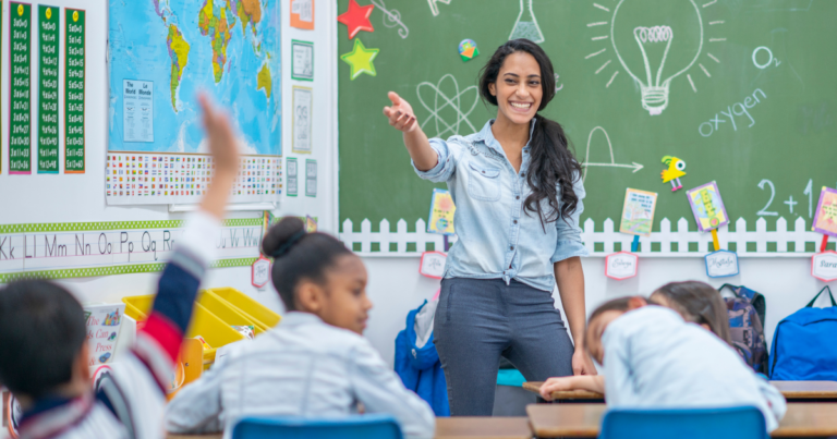 Logo for A female teacher standing in a classroom in front of a group of students